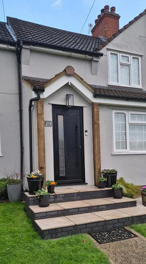 Front door of a house with a black door, pathway, and steps, surrounded by potted plants and shrubs.