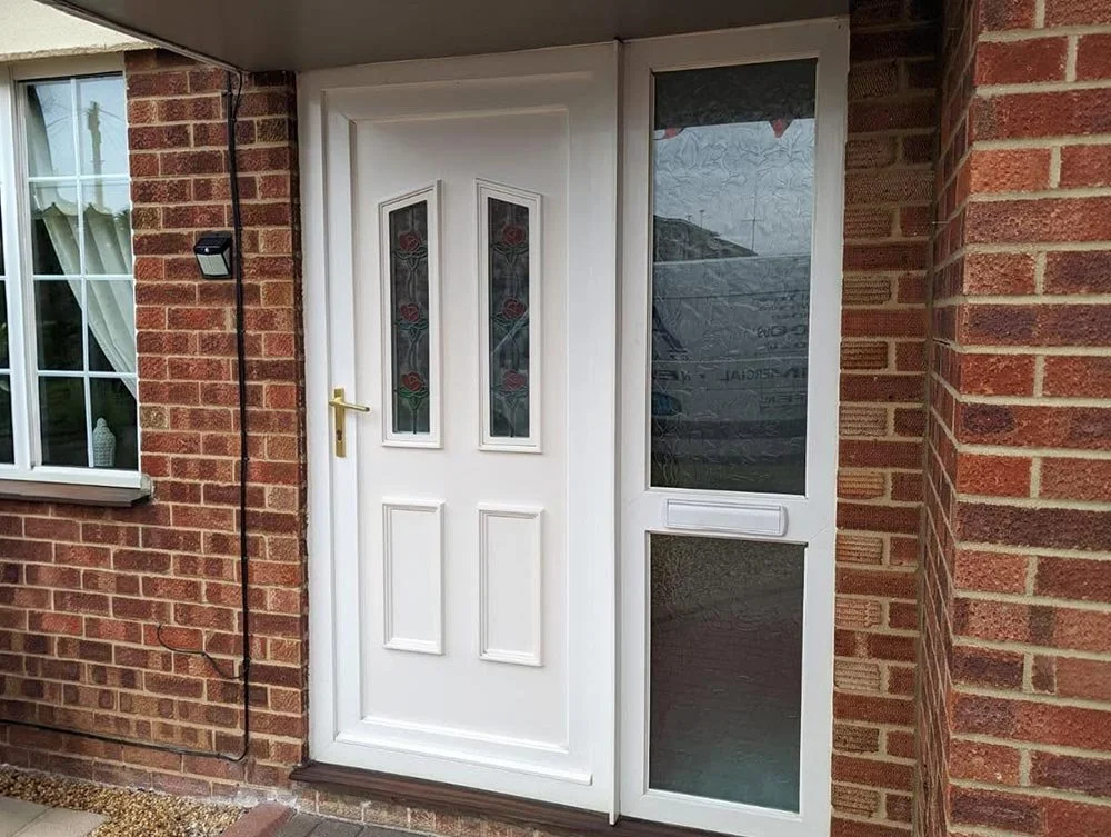 White front door with stained glass panels and a gold handle, surrounded by brick walls, with a window to the left and an enclosed porch with glass panels.