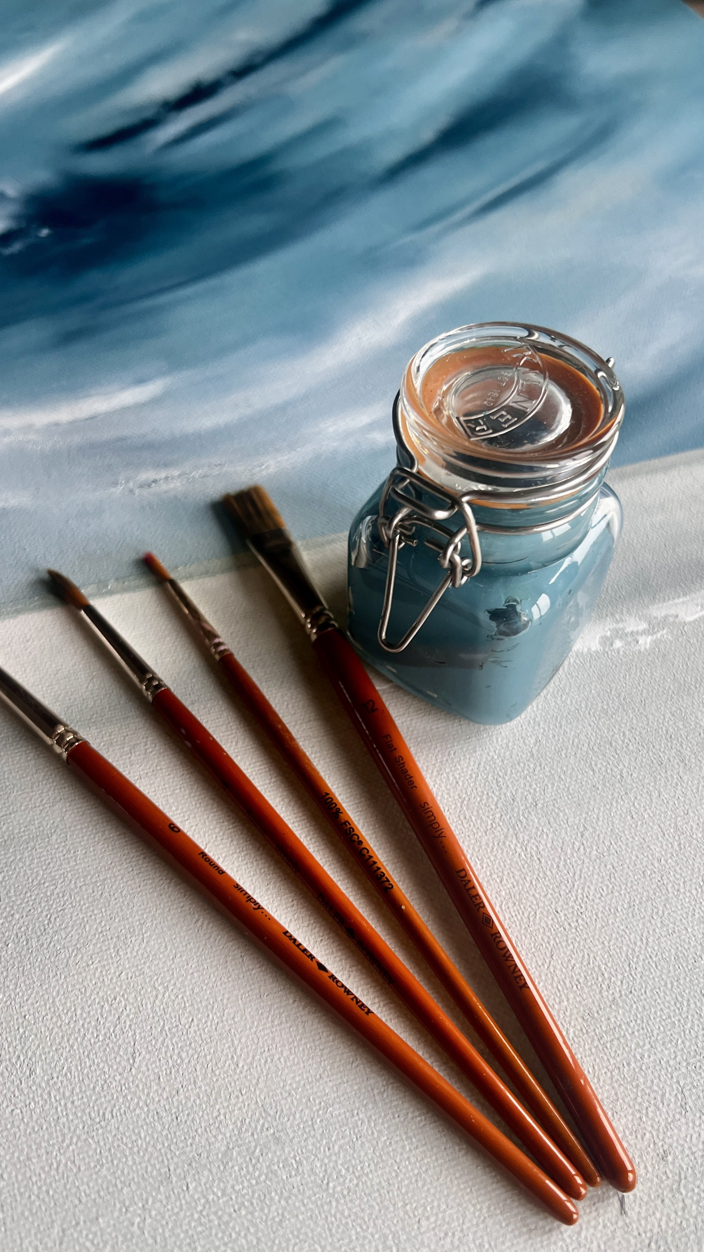 Four paintbrushes with orange handles, a small glass jar of brownish paint, and a blue abstract background.
