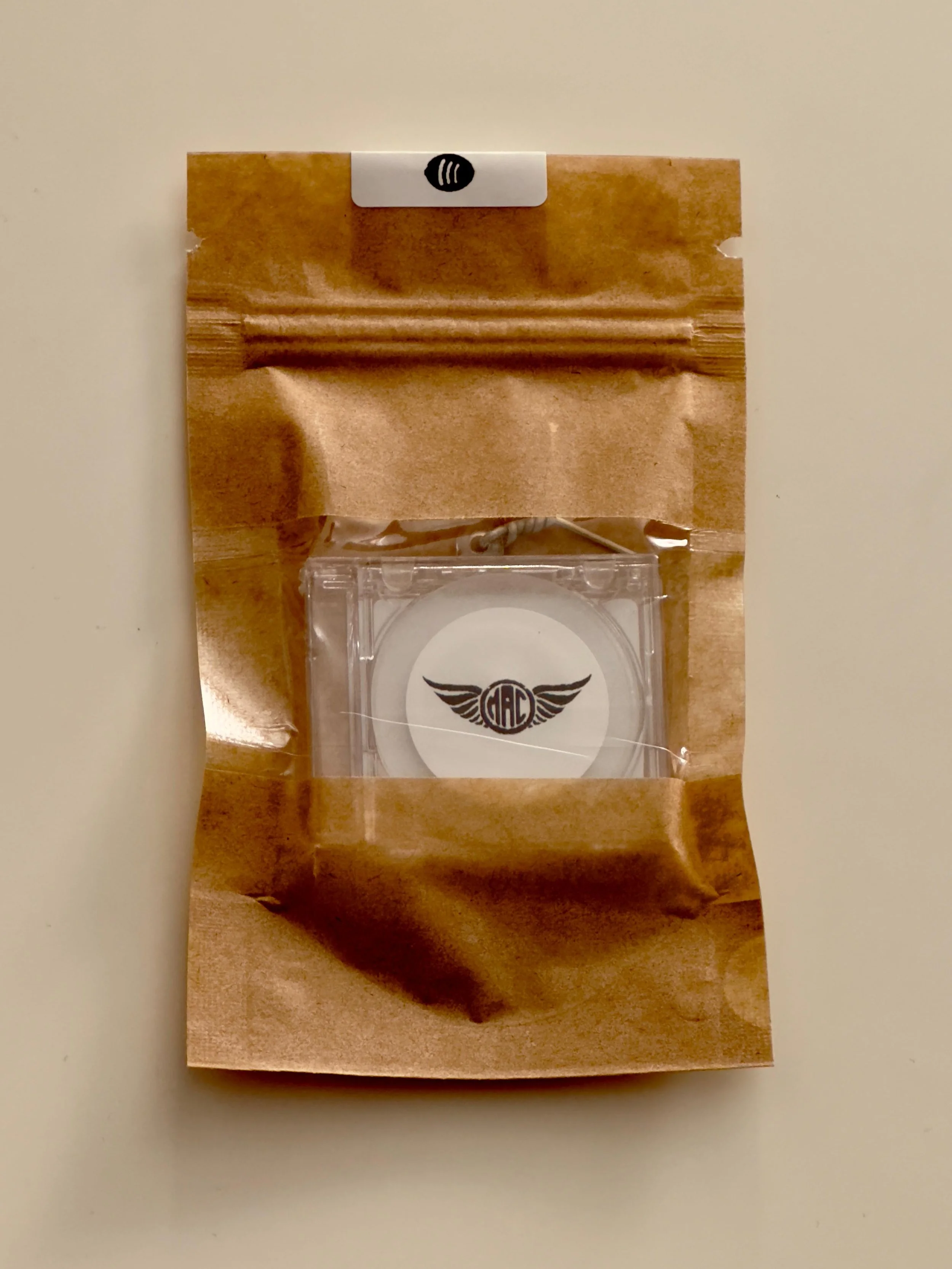 A sealed brown paper bag with a clear plastic window, containing a circular NFC object with a black logo featuring wings and the letters 'MAC', on a white surface.