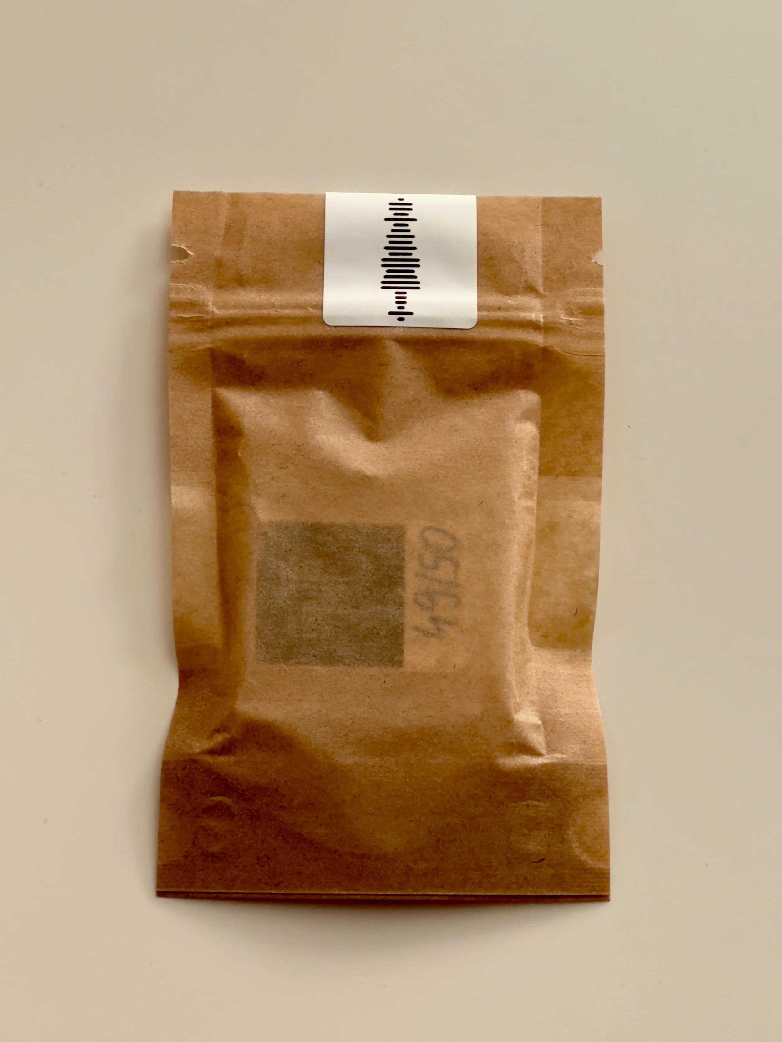The back of a brown paper bag with a transparent plastic window, containing a round object equipped with NFC technology, on which a physical seal has been affixed that also serves as a Spotify code.