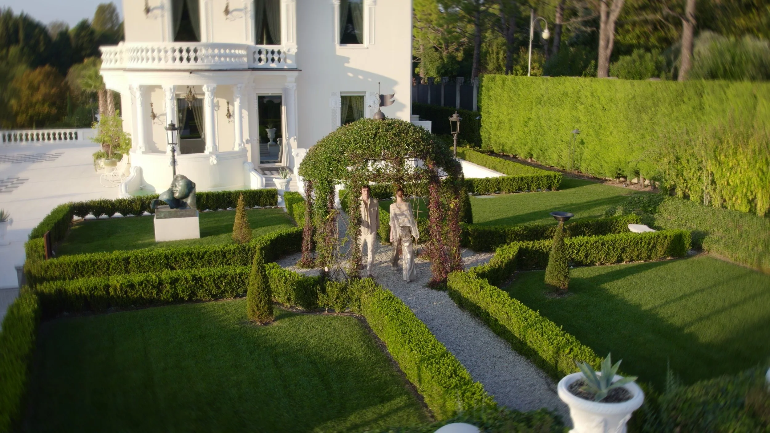 A couple walking on a gravel pathway through a well-manicured garden in front of a large white house with classical architectural details.