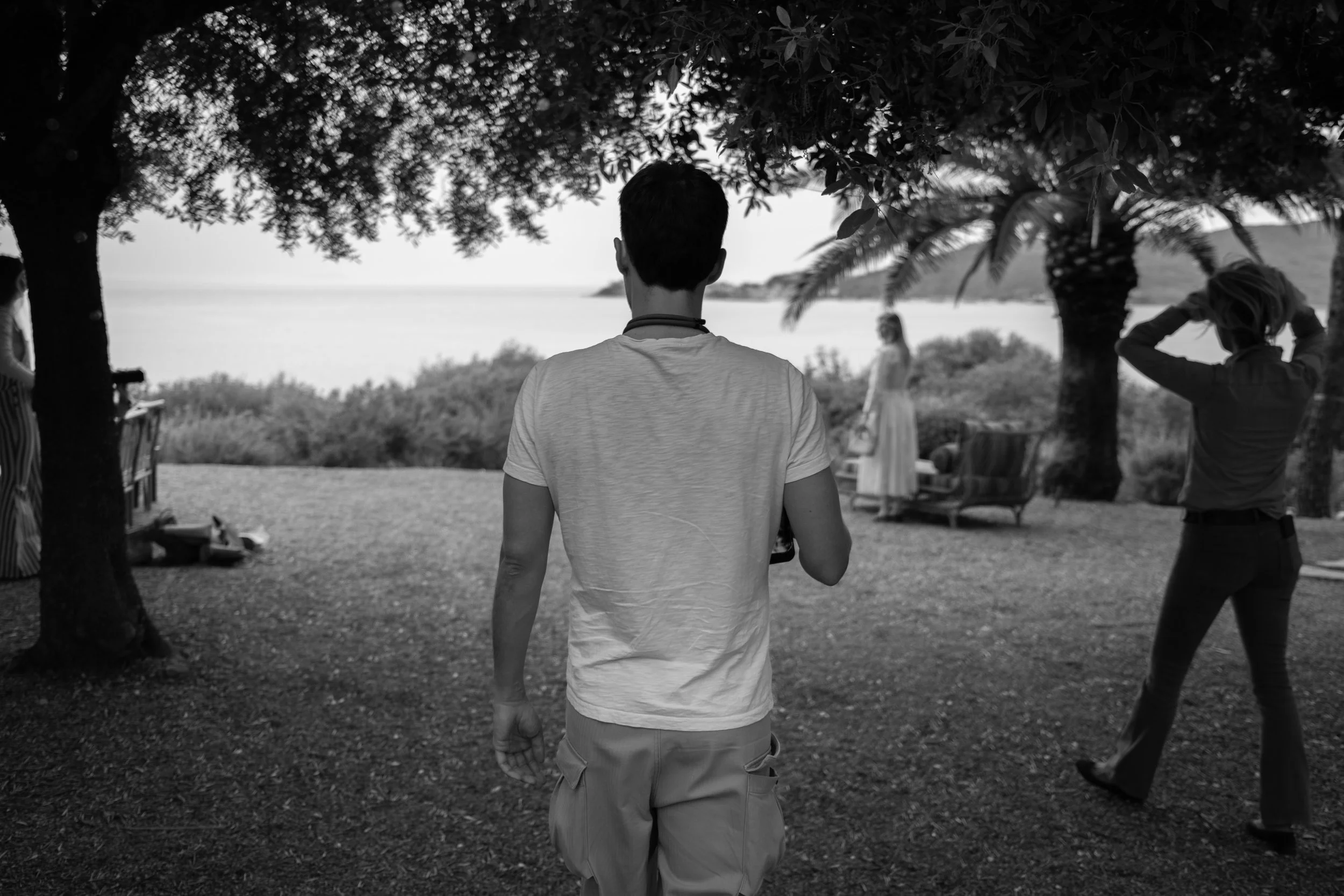 A black and white photo of a person with short hair, wearing a light-colored t-shirt and khaki shorts, standing outdoors under trees near a body of water. Two other people are visible in the background, one with hands on their head and the other standing near the water, possibly at a beach or lakeside.