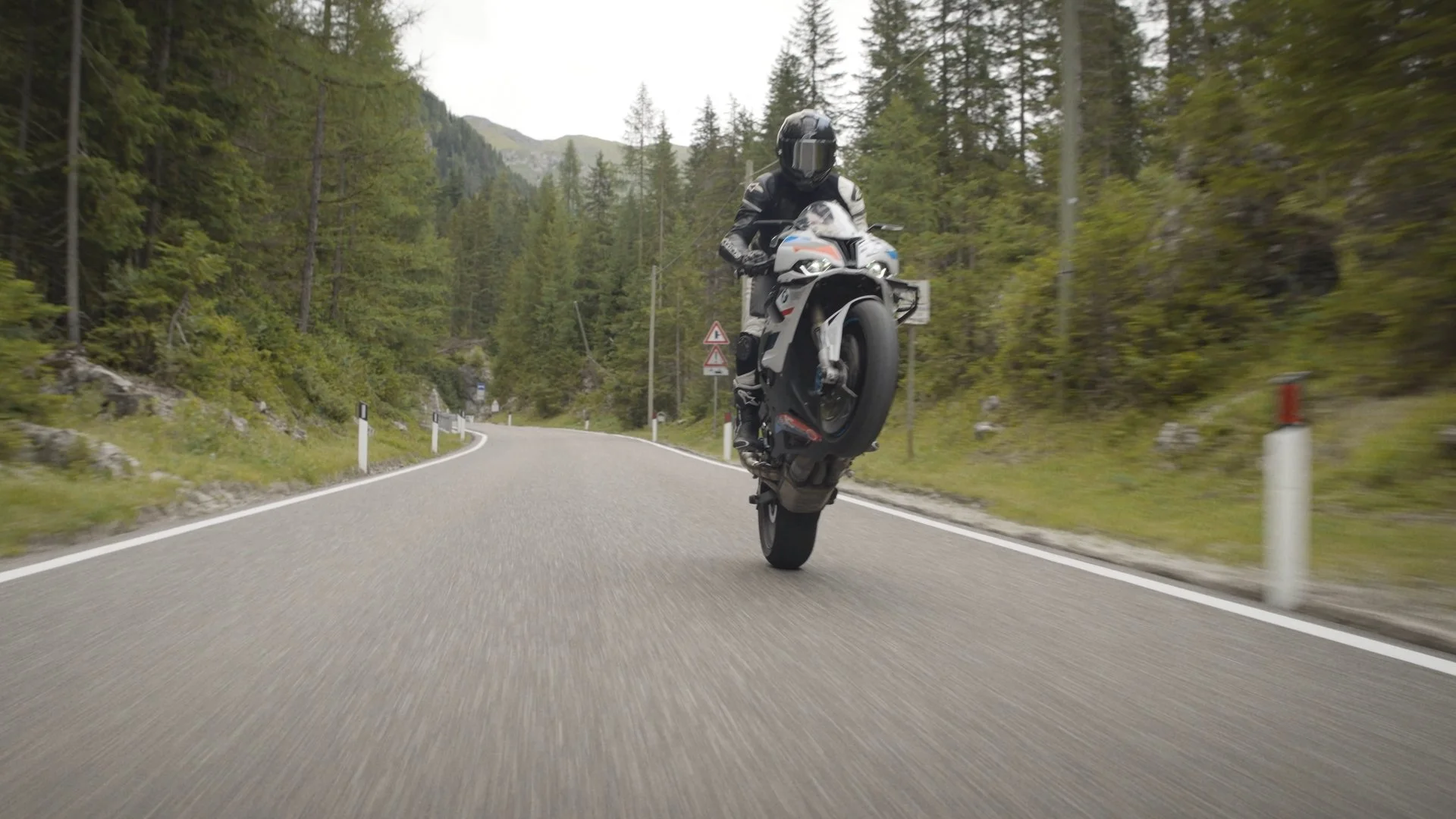 Motorcyclist performing a wheelie on a winding mountain road surrounded by trees.