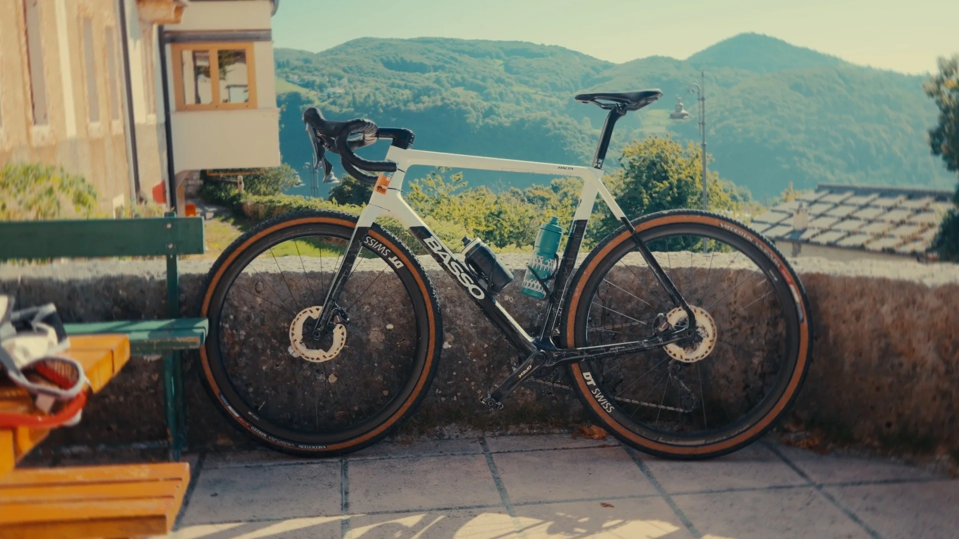A white and black mountain bike with brown tires leaning against a low concrete wall, with green hills and houses in the background.