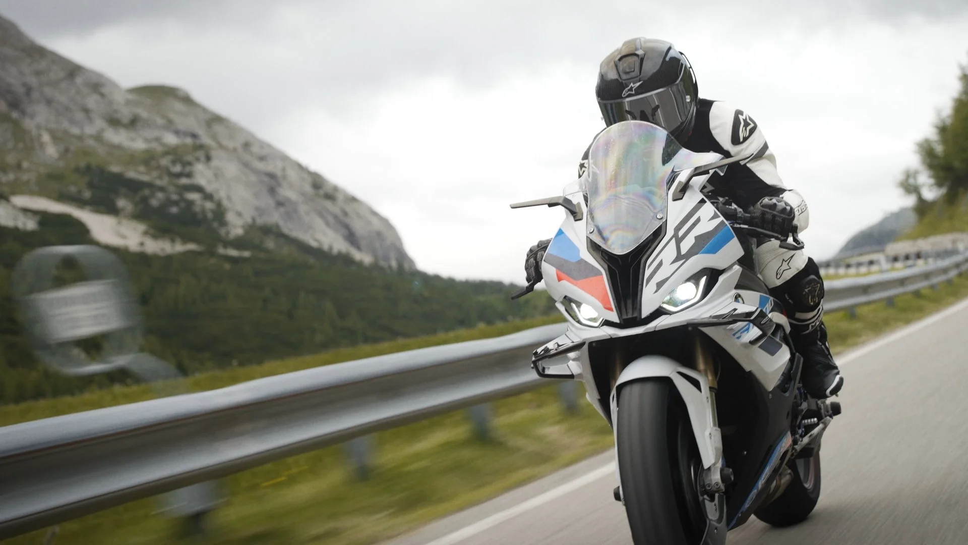 Motorcycle rider wearing a full helmet and protective gear riding a sports bike on a mountain highway with guardrail, surrounded by mountainous landscape and cloudy sky.