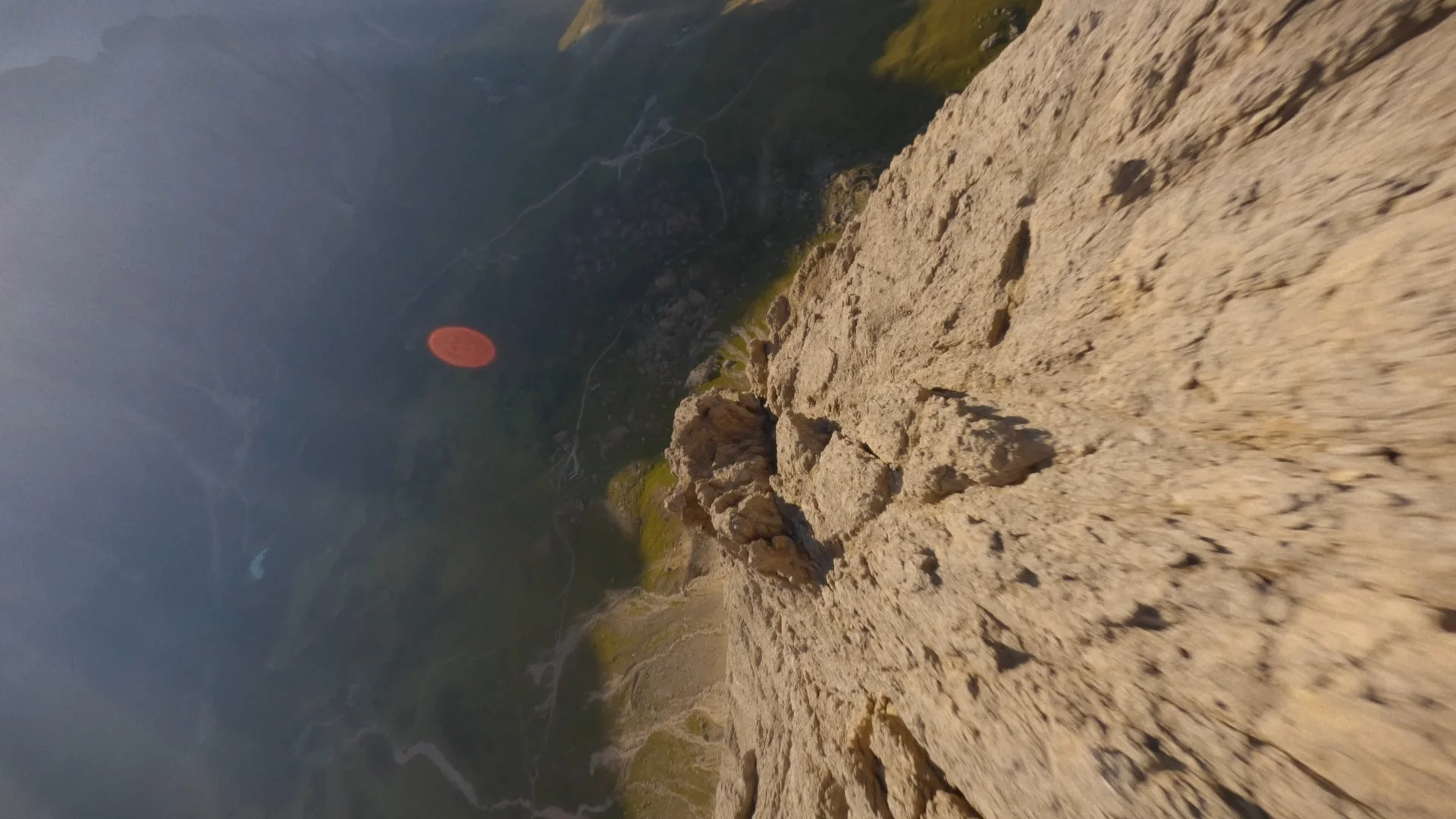The image shows a view from a steep, rocky mountain cliff looking down at the landscape below, including winding roads, green vegetation, and a distant horizon with a reddish sun or moon in the sky.