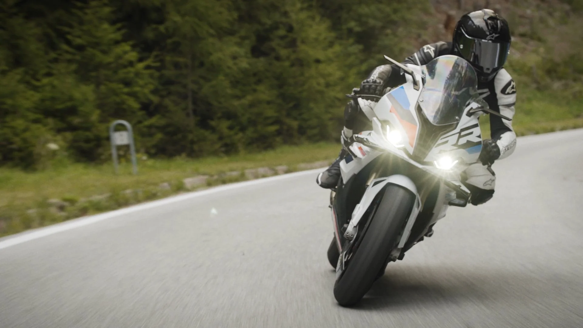 Motorcyclist wearing a black helmet and racing gear riding on a curving mountain road surrounded by greenery.