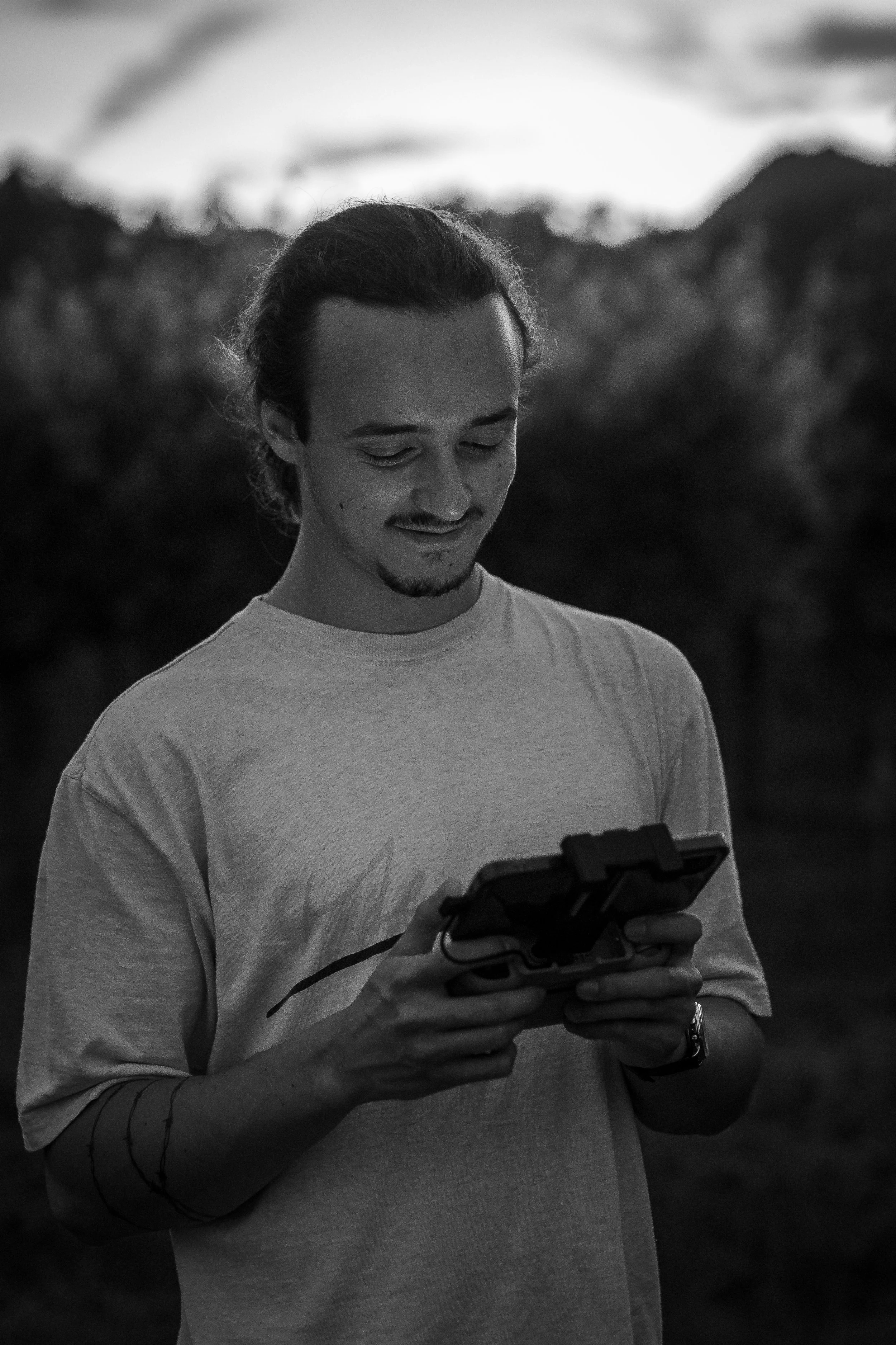A young man with ,wearing a light-colored T-shirt and wristwatch, is looking at a drone controller with a focused expression, outdoors in a natural setting.