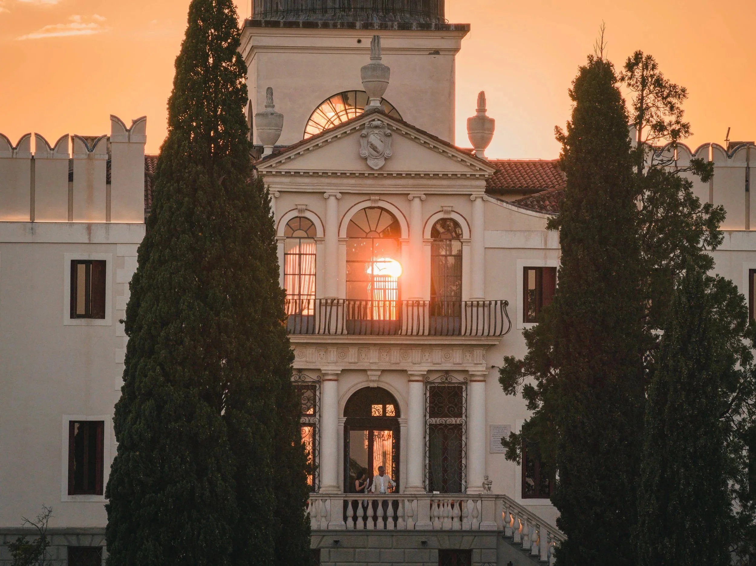 A historic white building with ornate architectural details and a clock tower, flanked by tall trees during sunset.