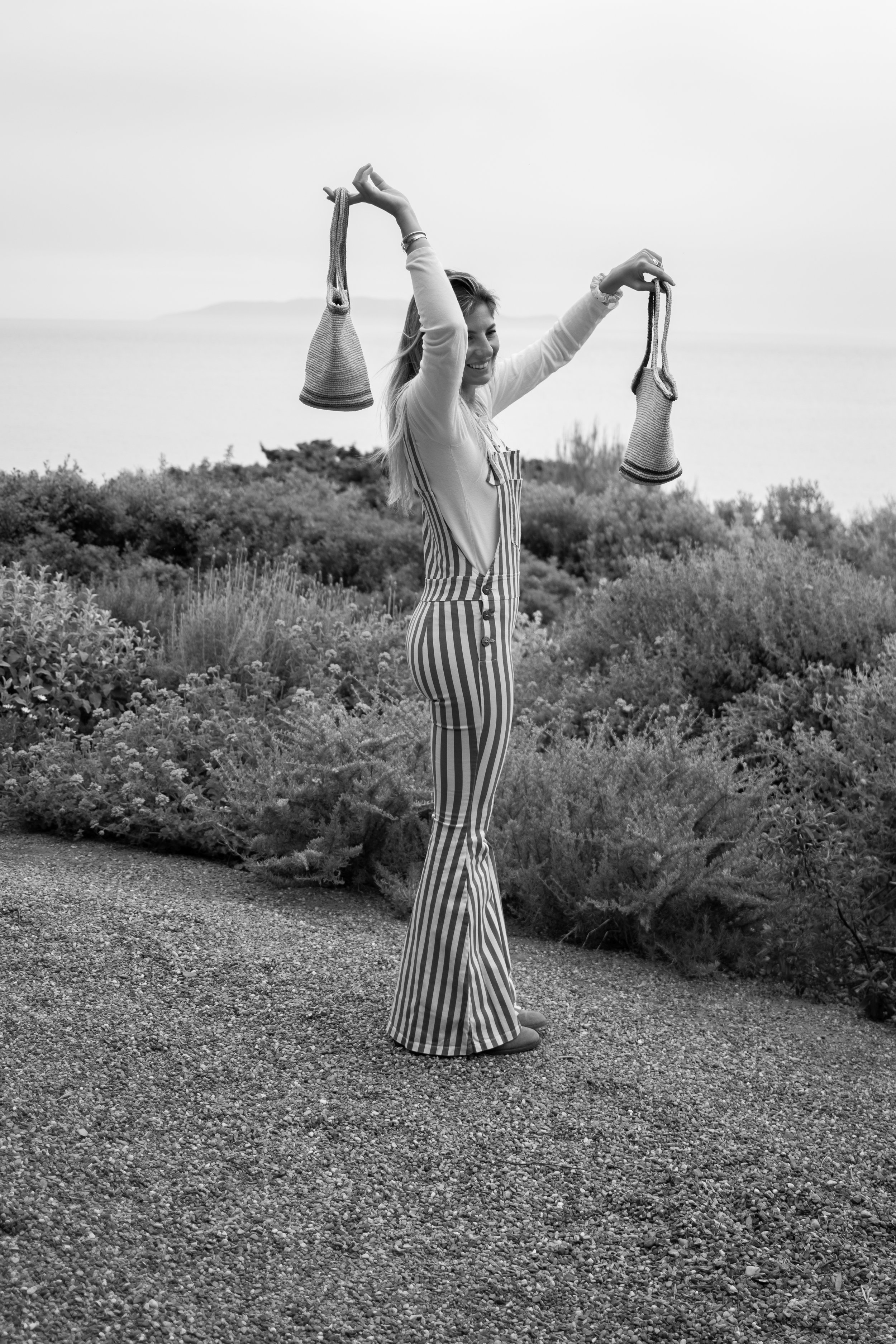 A woman standing outdoors near bushes by the beach, smiling, holding socks in each hand, wearing a light top and striped pants.