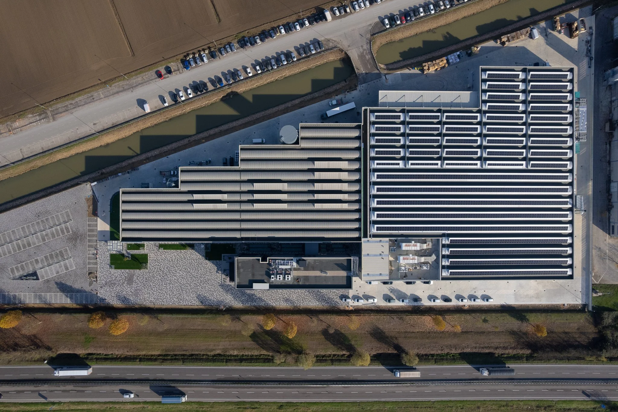 An aerial view of an industrial or commercial building with solar panels on the roof, adjacent to a parking lot with cars and trucks, separated by a canal or drainage ditch, with fields and a highway with moving vehicles nearby.