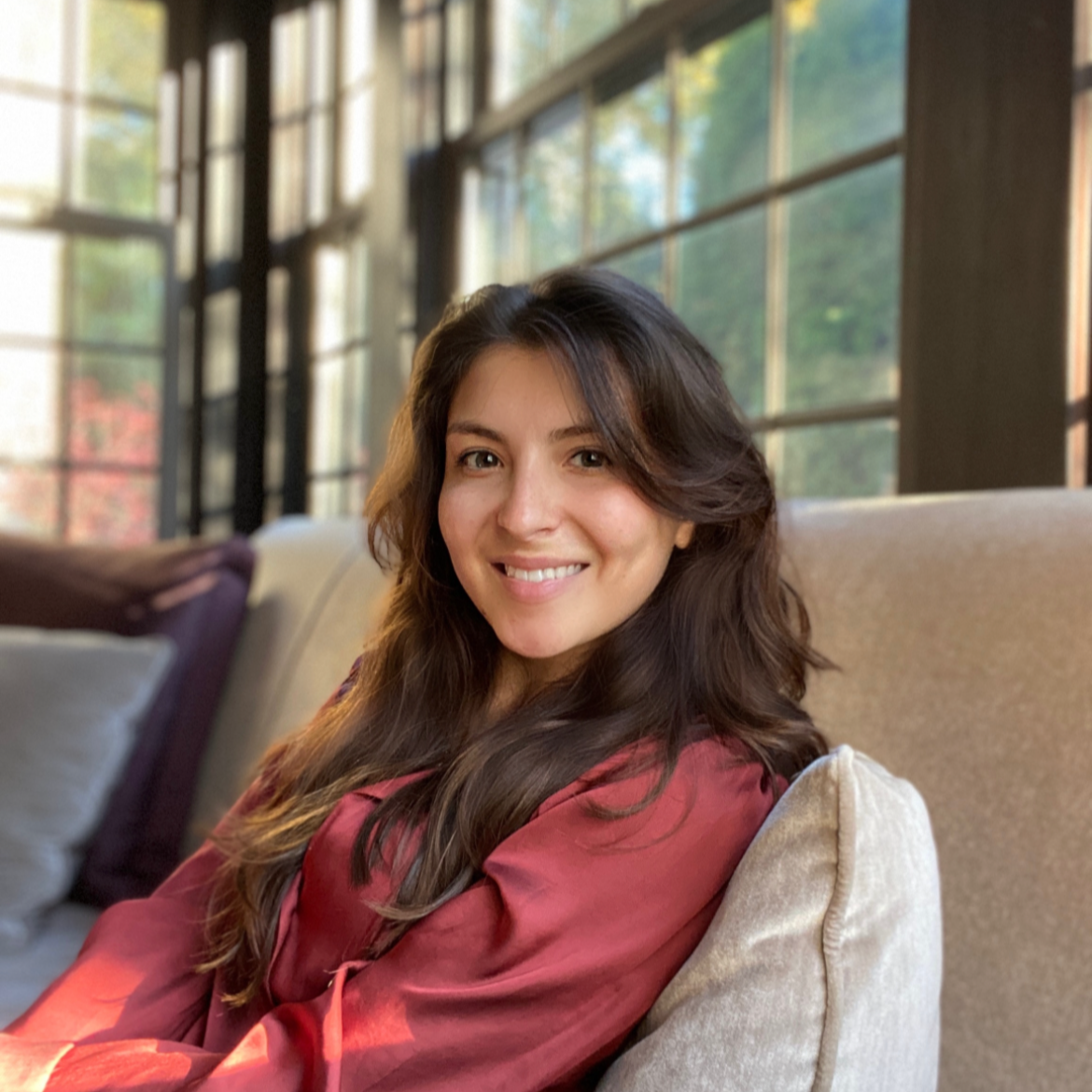 A woman with long, wavy brown hair smiling and looking at the camera, sitting on a beige couch in a sunlit room with large windows and greenery outside.
