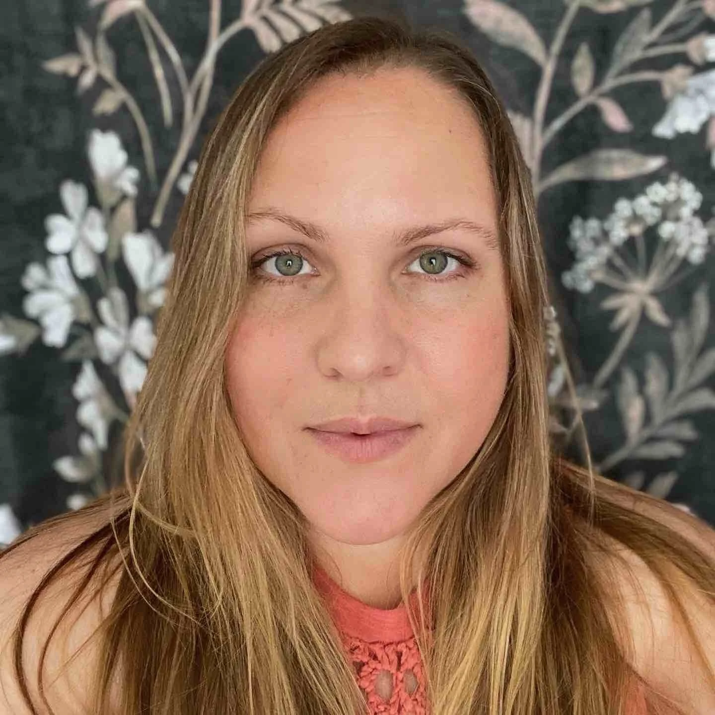 Close-up of EVA founder Darcey Rojas - a woman with long blonde hair, green eyes, wearing a pink top with a lace neckline. She is looking directly at the camera with a neutral expression, against a dark background with a floral pattern.