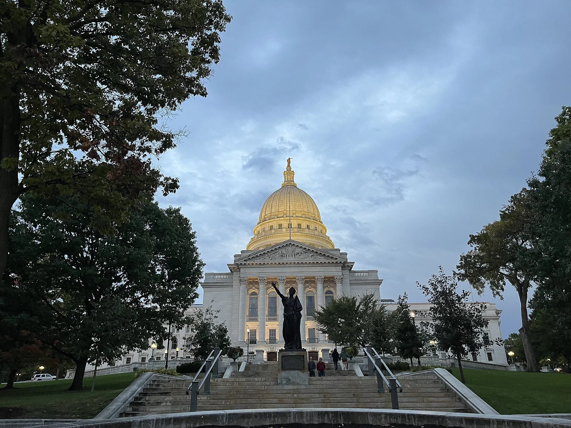 The image shows the state Capitol building in Harrisburg, Pennsylvania, with its large golden dome topped with a statue. The building is framed by trees on either side, and there are stairs leading up to the entrance with a statue of a woman holding a staff in the foreground.