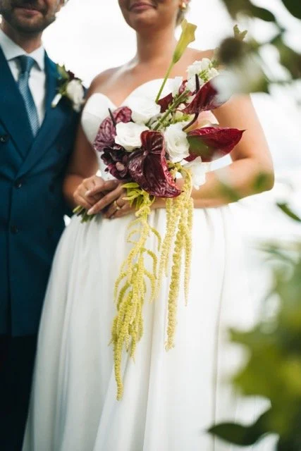 Brudparet håller i ett brudbukett med vita och röda blommor, omgiven av gröna blad, utomhus.
