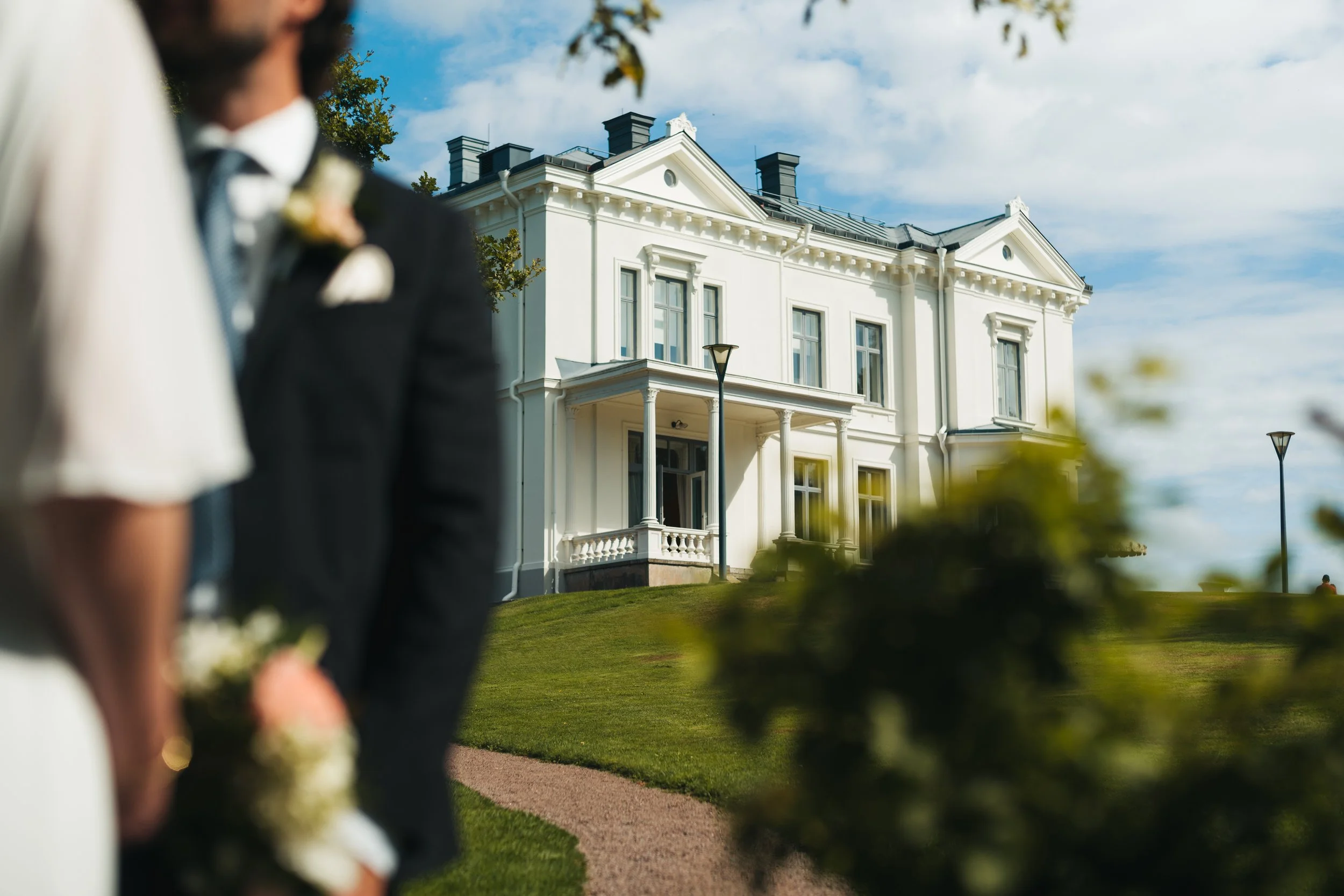En vit herrgård med kolonner och takutsmyckningar, under en blå himmel med få moln, sedd från en park där två personer står nära varandra i förgrunden, något utanför fokus.