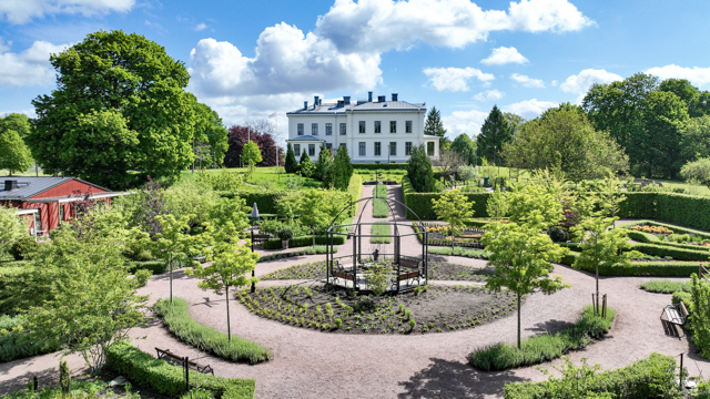 Vacker trädgård med en vit herrgård i bakgrunden, omgiven av gröna träd och blommor, under en blå himmel med moln.