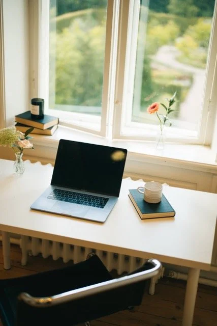 Ett skrivbord framför ett fönster med utsikt över grönska. På skrivbordet finns en öppen laptop, en kopp, ett anteckningsblock med en penna, och en vas med blommor.