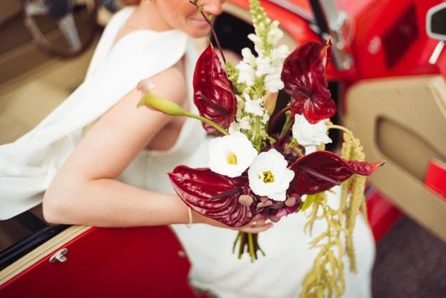 Person i vit klänning håller en bukett med vita, röda och gröna blommor inomhus.