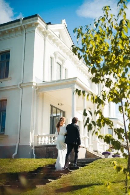Två personer i formella kläder står på trappan utanför en vit stor herrgård med tydliga klassiska detaljer, under en klar blå himmel, och ett träd med gula löv i förgrunden.