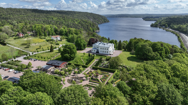 En stor herrgård med vita väggar är omgiven av en trädgård och grönskande natur nära en stor flod, med skog i bakgrunden.