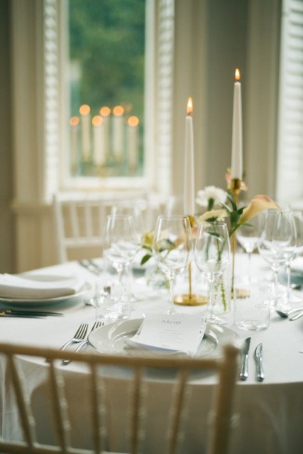 En elegant middagsborddukning med vita tallrikar, glas, bestick, blomsterarrangemang och två tändstickor i ljuslyktor i ett rum med stora fönster.