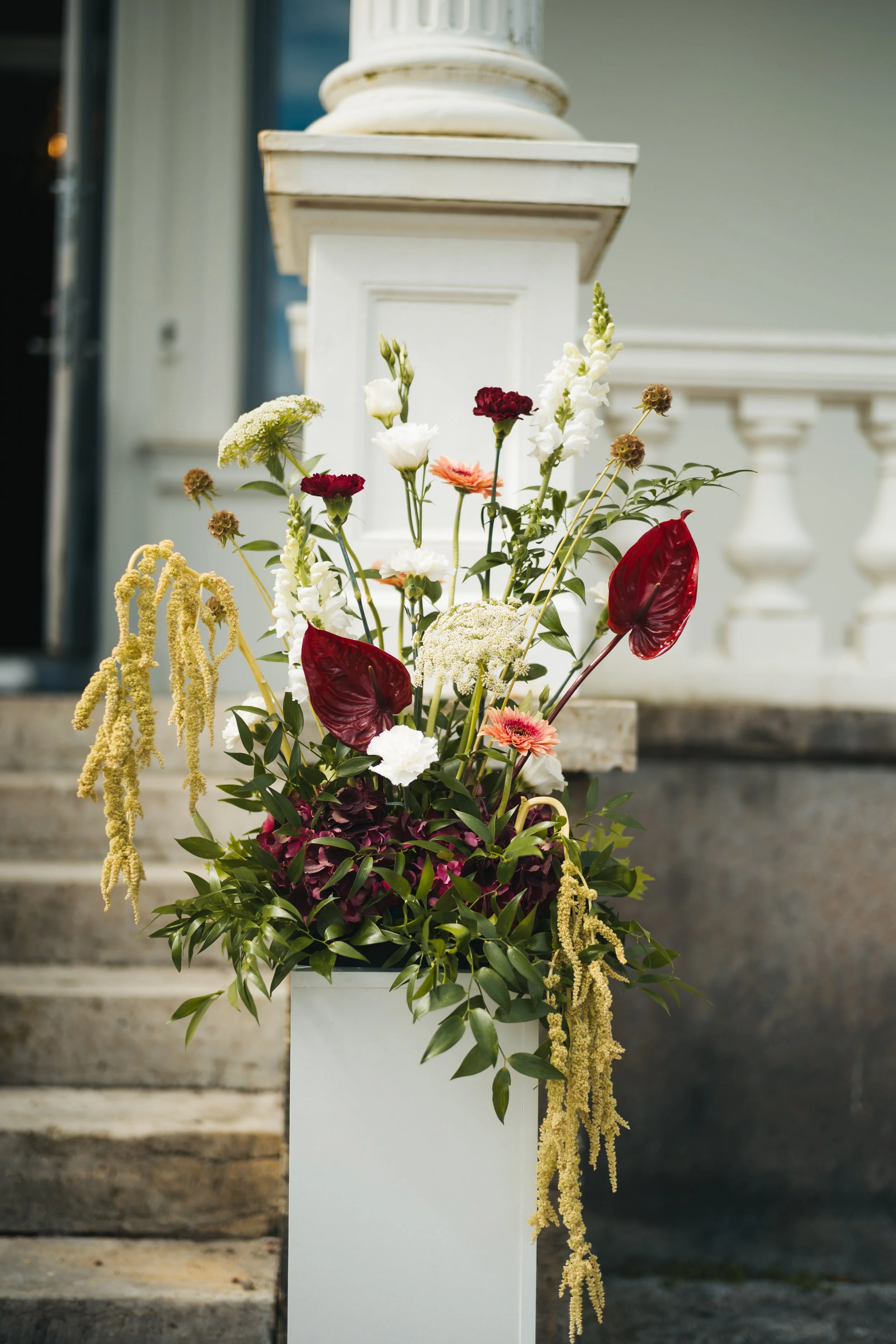 Färgglad blomsterarrangemang med olika sorters blommor, placerad framför en vit pelare på trappsteg.
