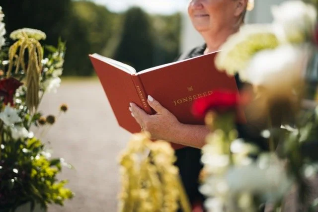 En äldre kvinna som ler och läser en röd bok utomhus bland blommor.