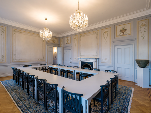 Ett elegant mötesrum med vita väggar med guldaccenter och ett eldstadsparti, två kristallkronor, ett stort långt bord med svarta stolar, och ett klassiskt persiskt matta.