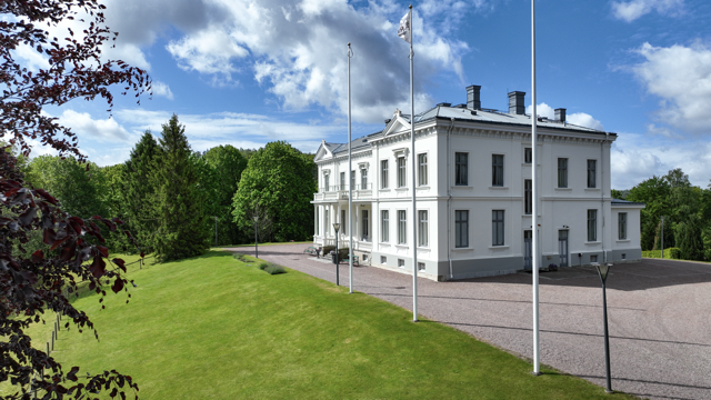 En vit herrgård omgivet av gröna träd och en gräsmatta, med tre flaggstänger framför byggnaden under en molnig himmel.