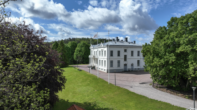 En vit byggnad omgiven av gröna träd och en gräsyta, med en blå himmel och moln ovan.