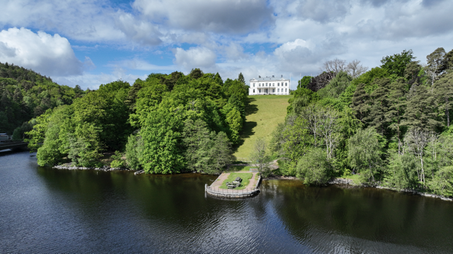 Landskap med sjö, gröna träd och en vit stor byggnad uppe på en kulle under en molnig himmel.