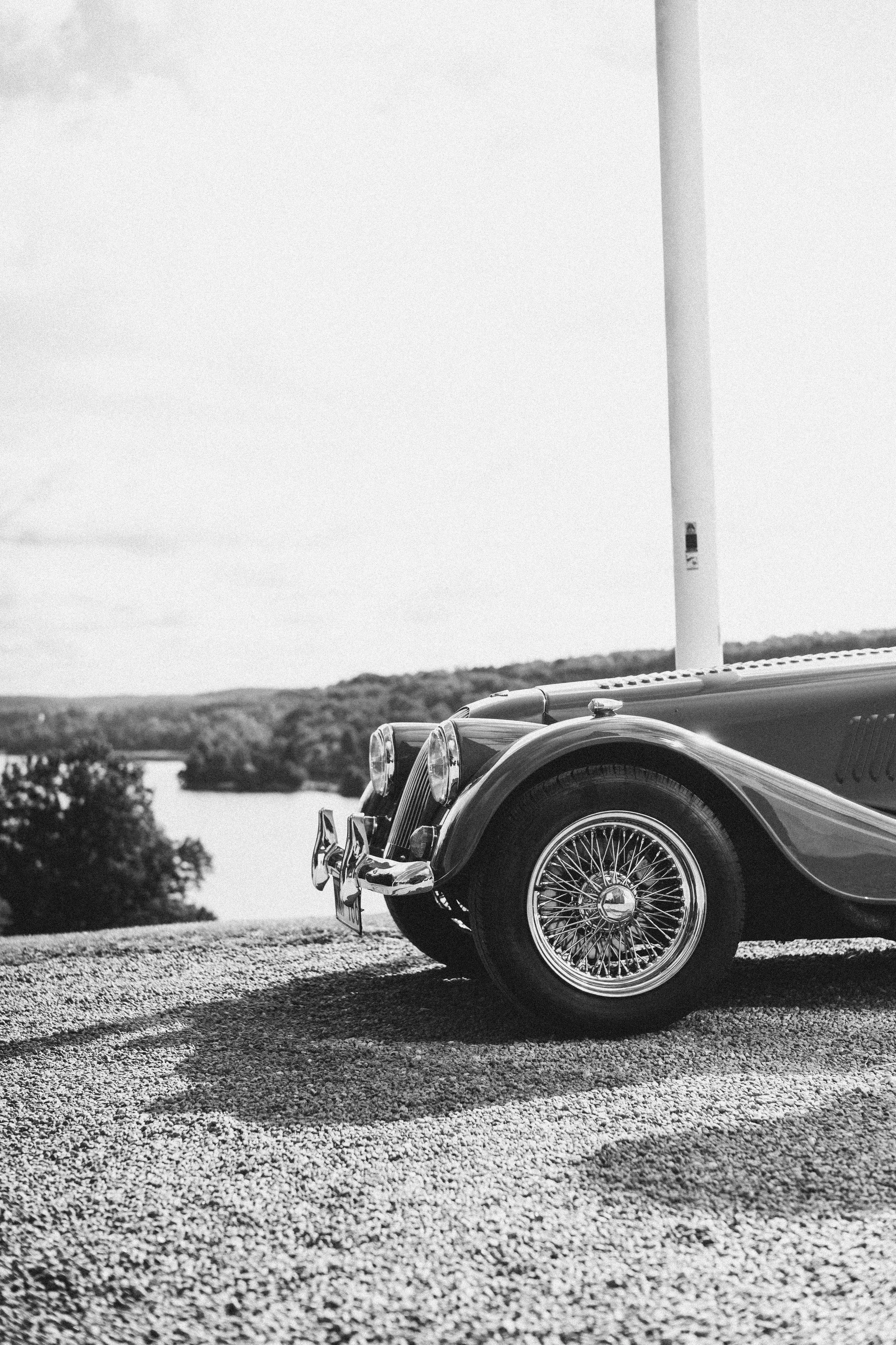 En svart vintage bil med chrome detaljer och spindeltrådshjul, parkerad på en grusväg med landskap i bakgrunden.