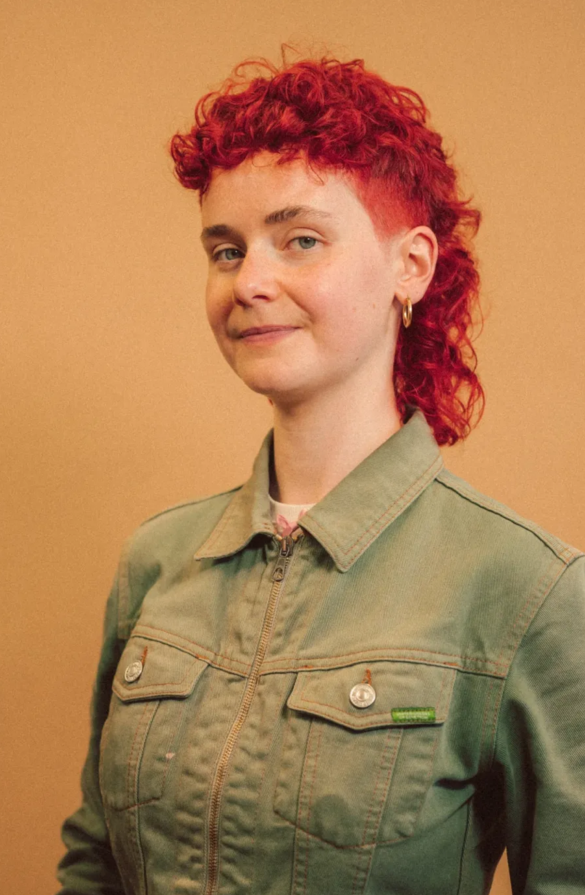 A woman with short, curly red hair and piercing blue eyes, wearing a light green denim jacket with metal buttons, standing against a beige background.