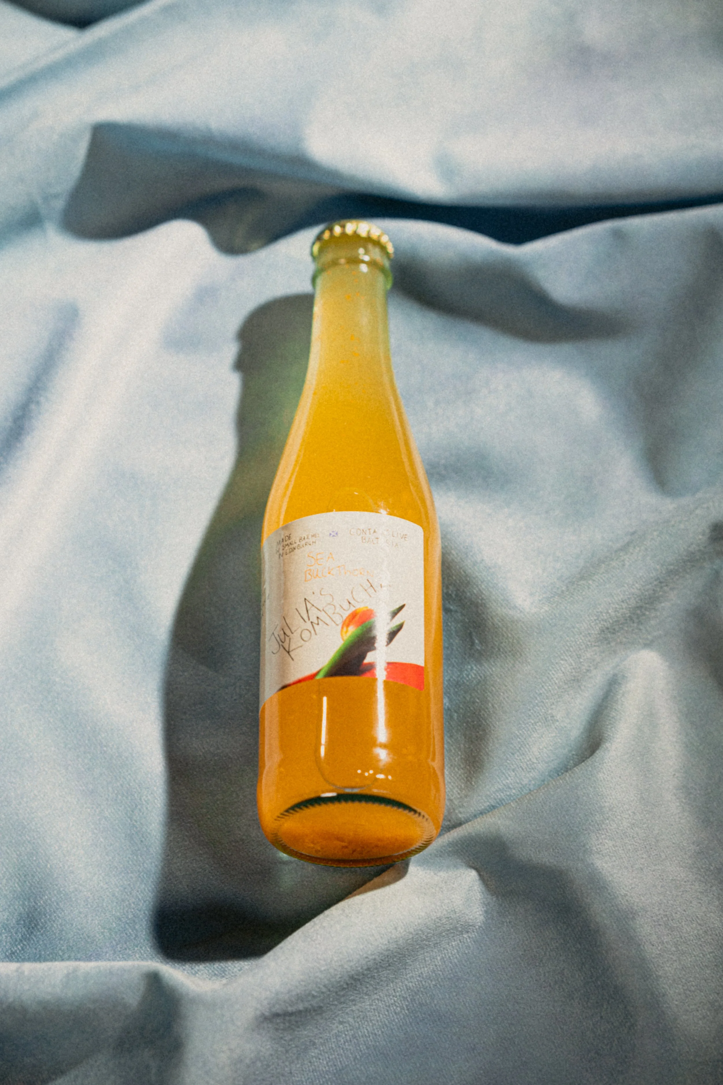 A glass bottle of orange juice with a label that has a rainbow graphic and some handwritten text, placed on a light blue satin fabric.