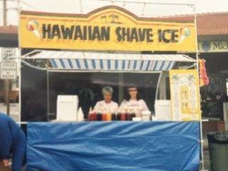 Two women standing behind a street vendor stand with a yellow sign that reads 'Hawaiian Shave Ice'.