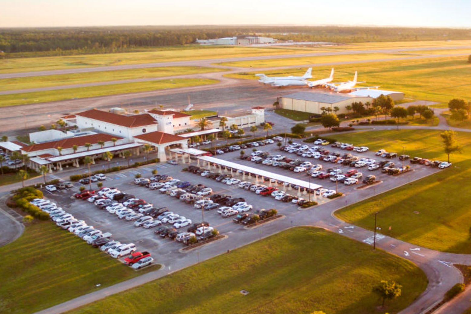 Aerial view of an Brunswick Golden Isles Regional Airport, located in Brunswick Georgia, with dotted lines of parked vehicles and three private jets on the runway at sunset.