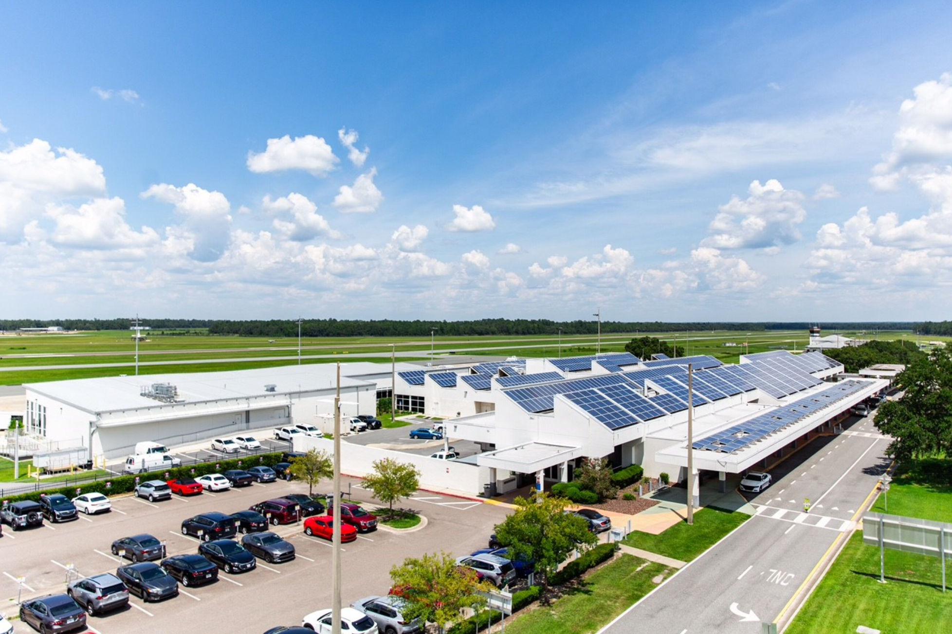 Aerial view of a commercial building with solar panels on the roof. Parked cars are visible in the parking lot, with a green field and cloudy sky in the background.