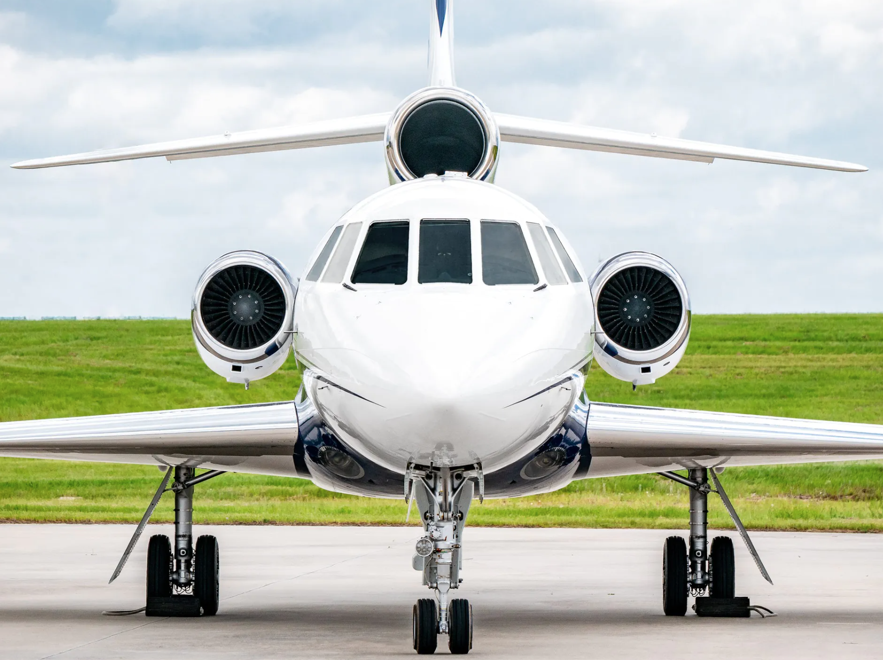 Front view of a white private jet on a tarmac with grass and cloudy sky in the background.