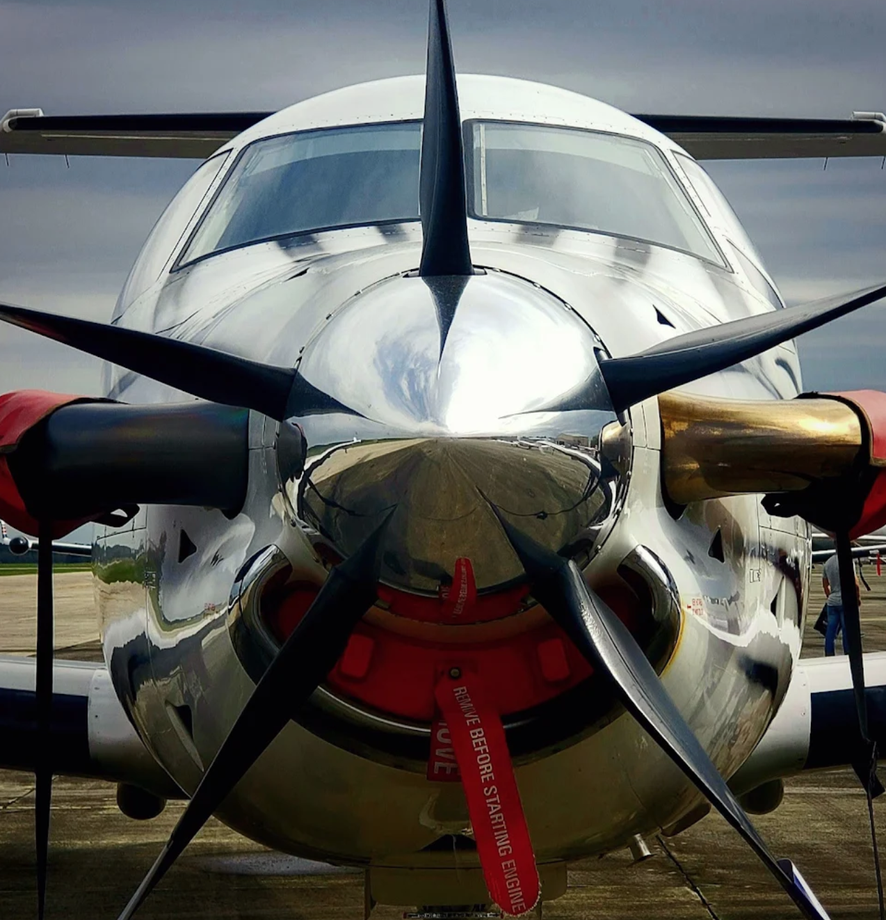 Close-up front view of a propeller plane with a reflective metallic surface, showing its nose, propellers, and cockpit windows. The airplane is parked on a runway under a cloudy sky.