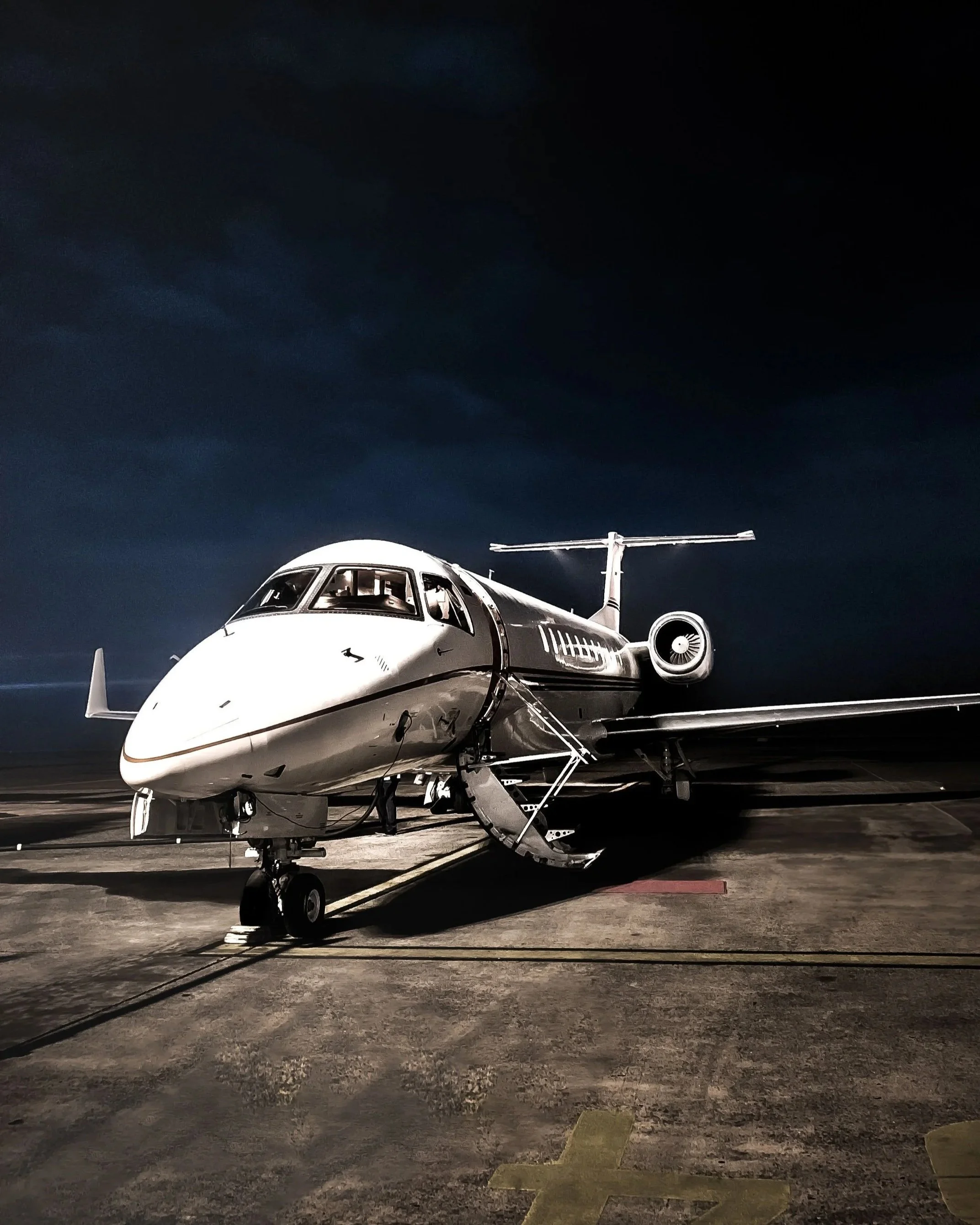 Nighttime view of a white private jet parked on tarmac with a helicopter tail visible in the background, under dark sky.