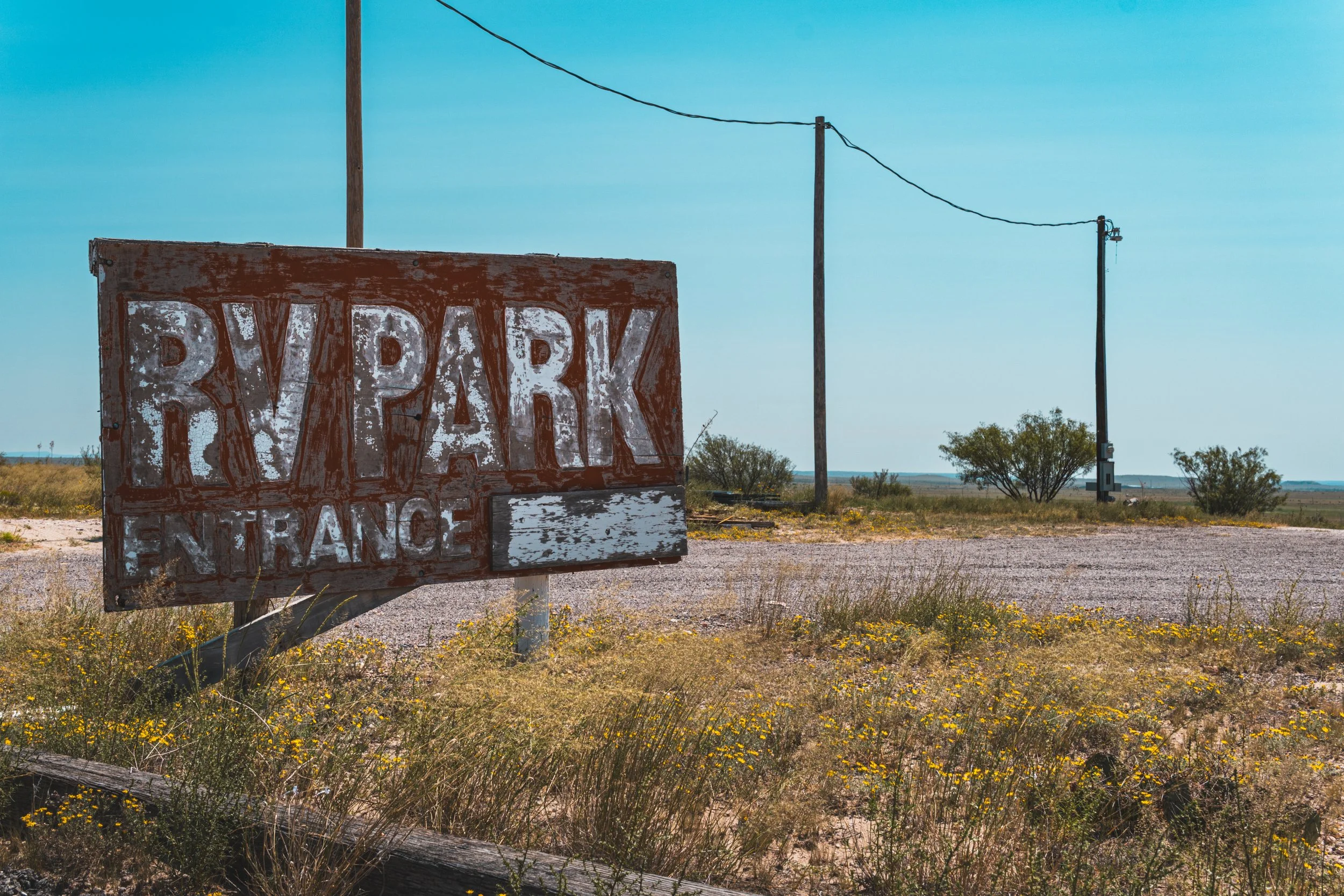 A weathered wooden sign that reads 'Riverrun Ranch' with the word 'Ranch' partly obscured, situated in a rural landscape with power poles, trees, and a clear blue sky in the background.