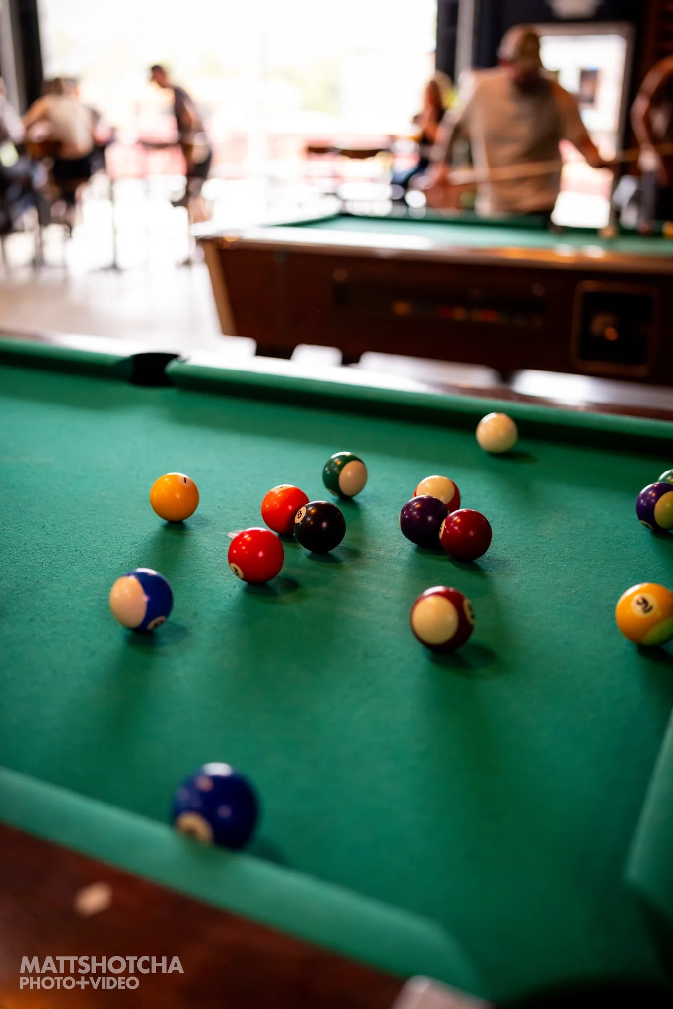 Close-up of a pool table with numbered billiard balls scattered across the green surface, with a blurred background of people at The Burl in Lexington.