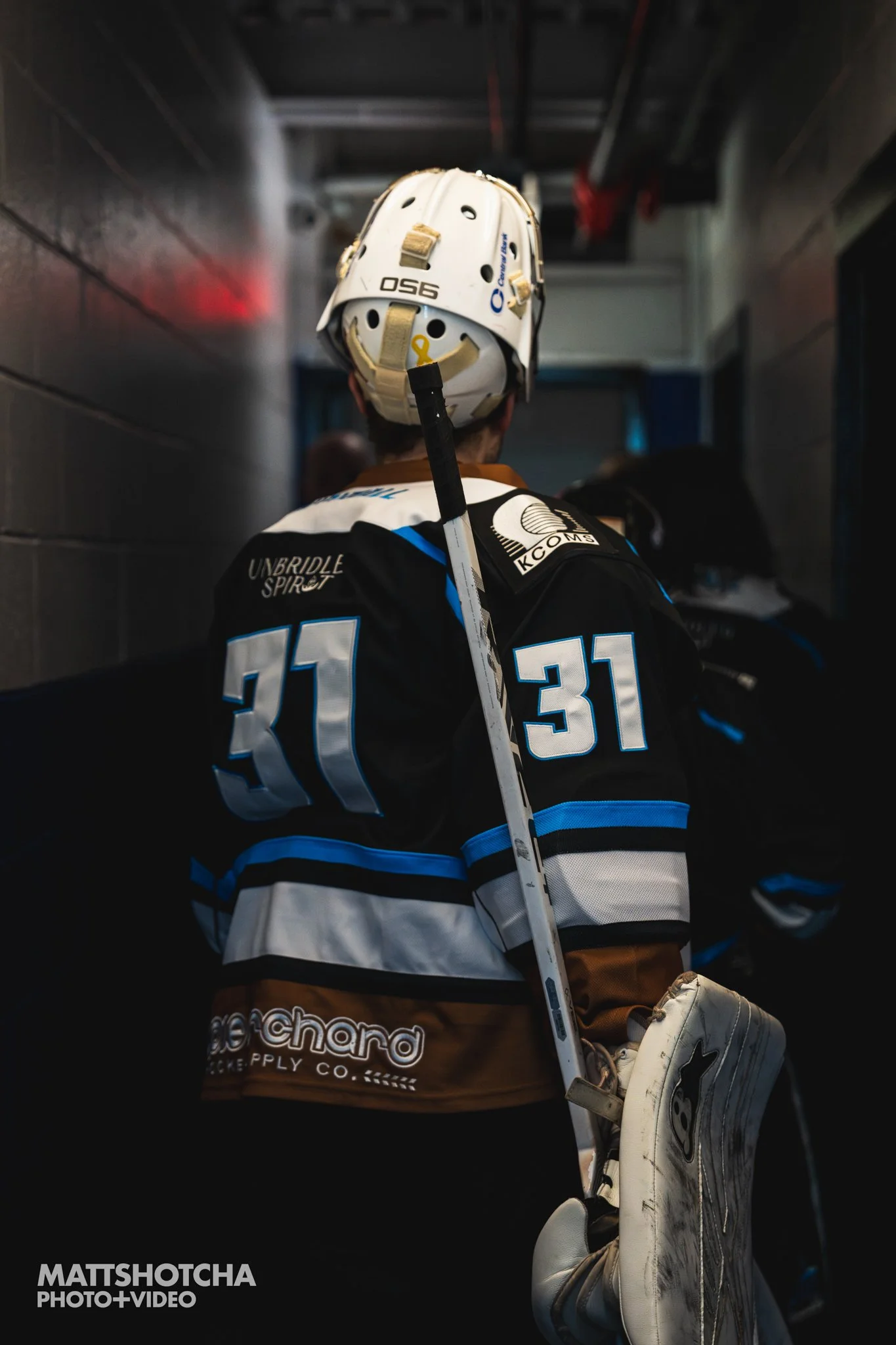 Back of a Lexington Nightfall hockey goalie in black, blue, white, and brown jersey holding a hockey stick, with a hockey glove and blocker in the foreground. The background shows the locker room tunnel.