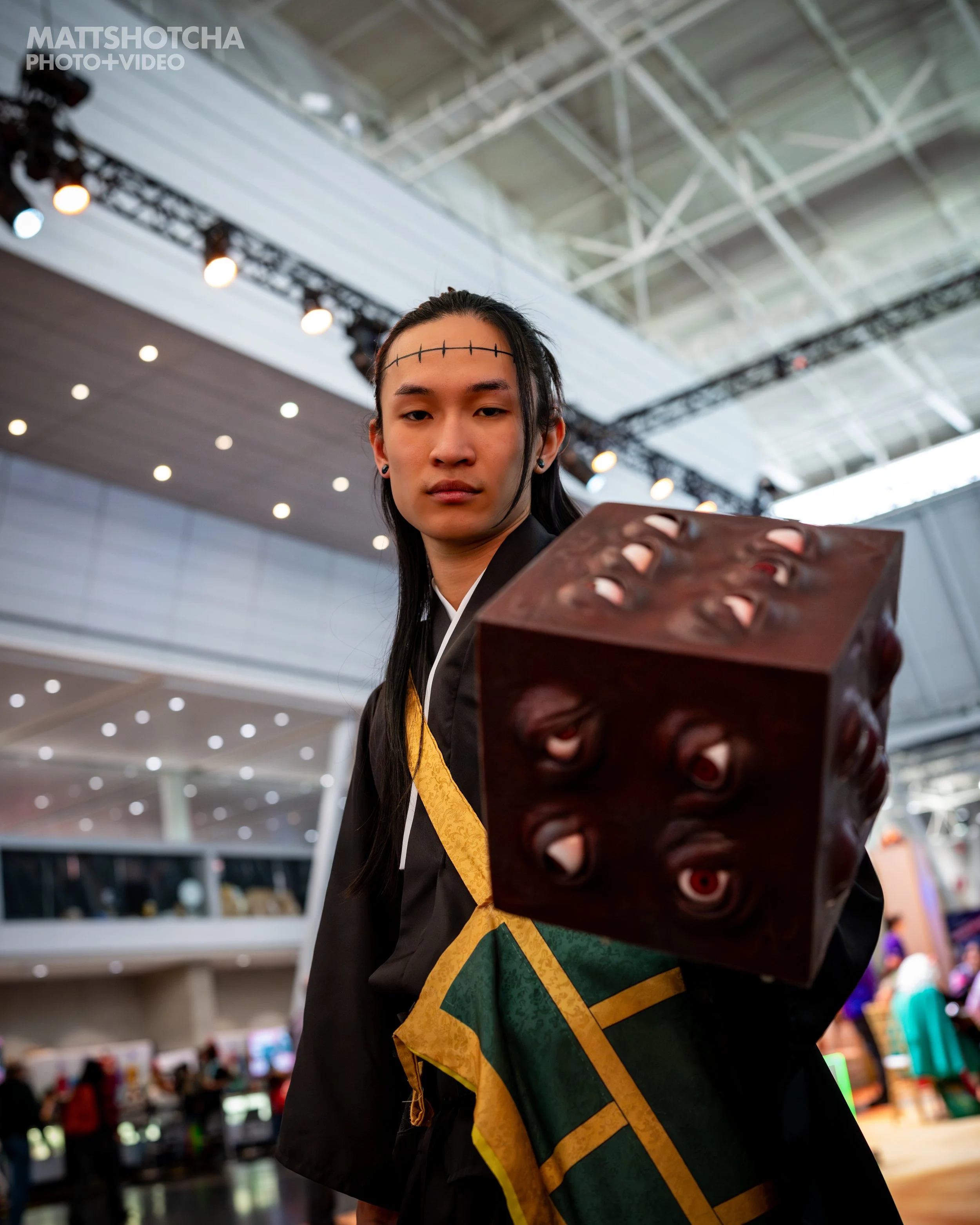 A man, Derailed3d, dressed as Kenjaku from Jujutsu Kaisen, holding a prop Prison Realm with multiple eyes, standing in a brightly lit indoor space at PAX East 2026.