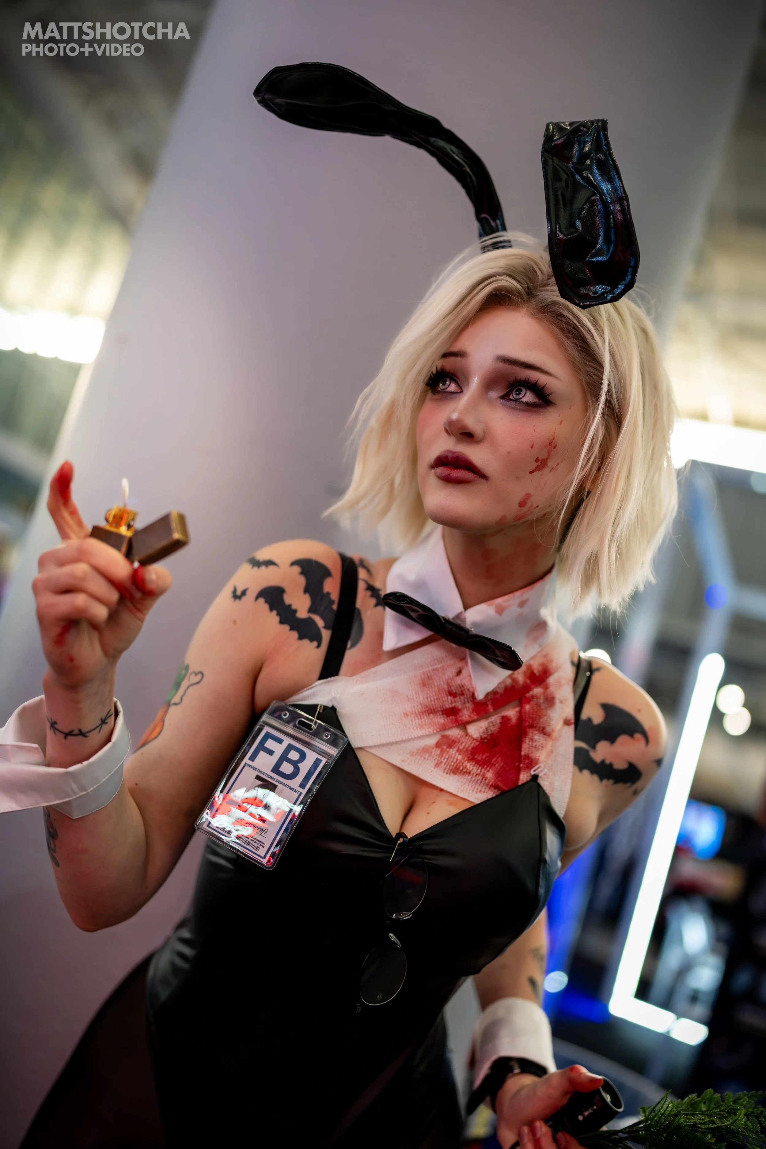 A woman, RoseWaterCosplay, dressed as Bunny Grace from Resident Evil with black bunny ears, a blood-stained white shirt, and fake blood on her face and arms, holding a cigarette lighter and an 'FBI' badge around her neck.