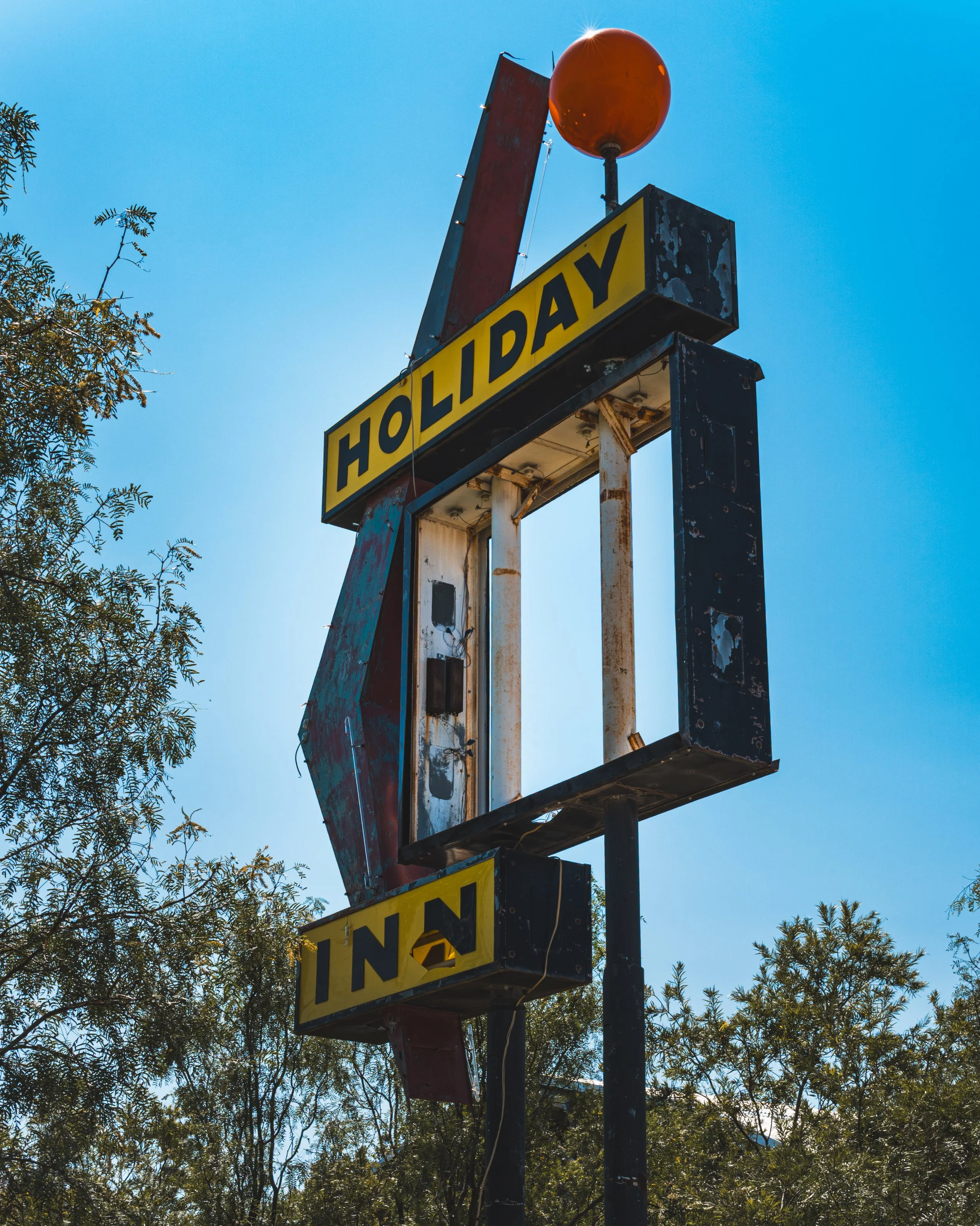 Old, rusty billboard frame with a yellow sign that says 'HOLIDAY INN' and a large orange spherical light on top against a clear blue sky, with trees surrounding the structure.