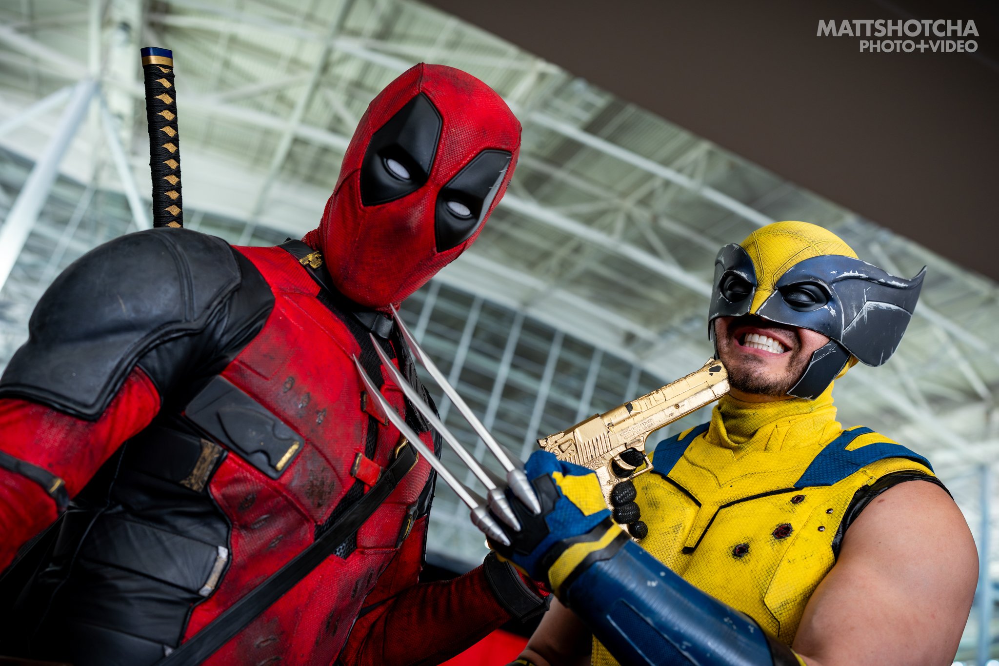 Costumed individuals dressed as Deadpool and Wolverine, holding guns, posing in a modern indoor setting with a glass and metal ceiling.