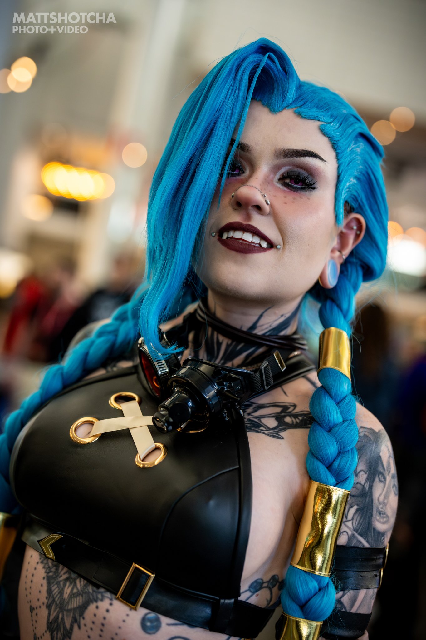 A woman, HoloFox69, dressed as Jinx from League of Legends. She is wearing a black outfit with gold accents and a black harness with gold loops. She has a confident expression on her face.